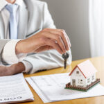 person-holding-house-keys-toy-model-house-desk